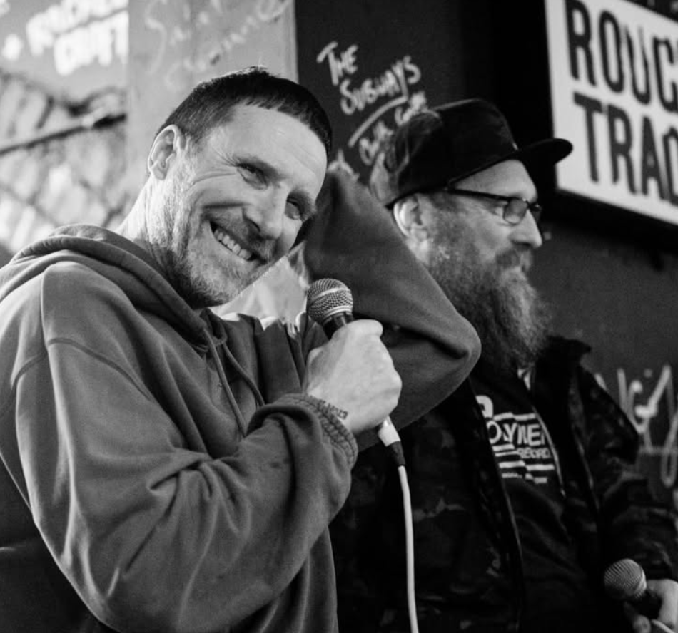 Sleaford Mods smiling outside Rough Trade, one holding a mic, in black and white