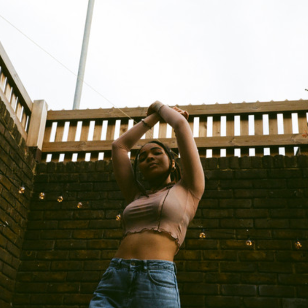 Chinwe posing against a brick wall and wooden fence, arms raised above her head