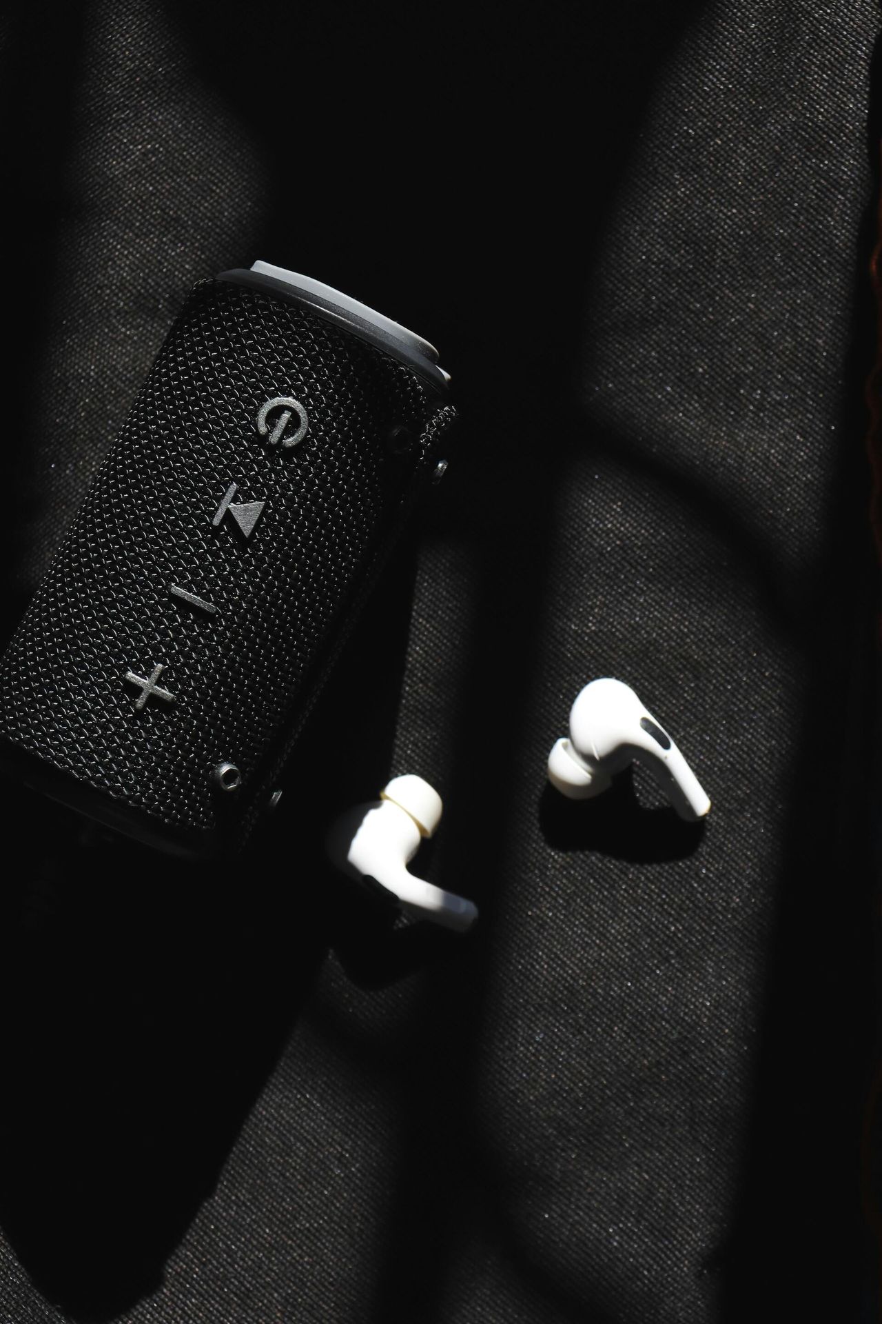 Black portable speaker and white wireless earbuds on dark fabric