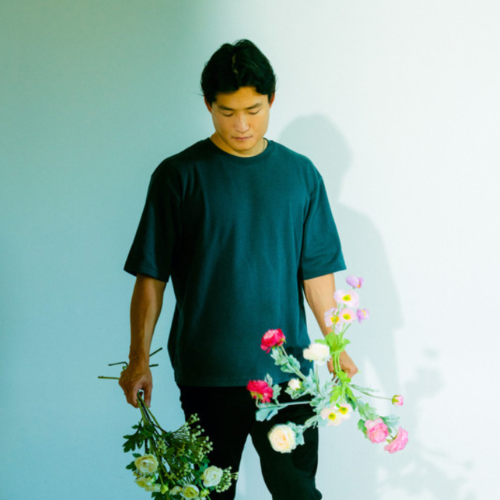 Young man in a green T-shirt holding two bunches of flowers against a pale wall
