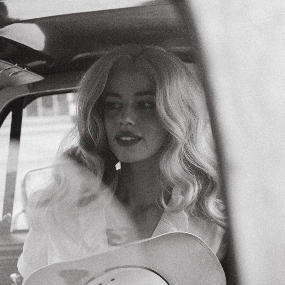 Woman with long blonde waves in a vintage car holding a cowboy hat, black and white