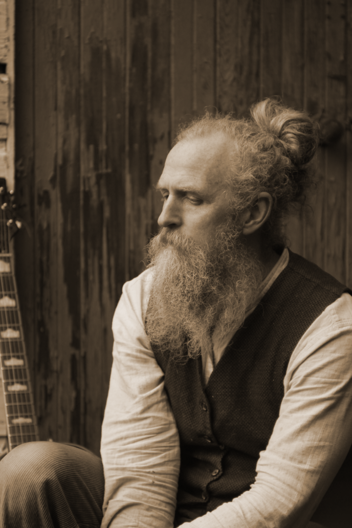 Brick Turpin in a sepia portrait, long beard and waistcoat, seated by a wooden wall
