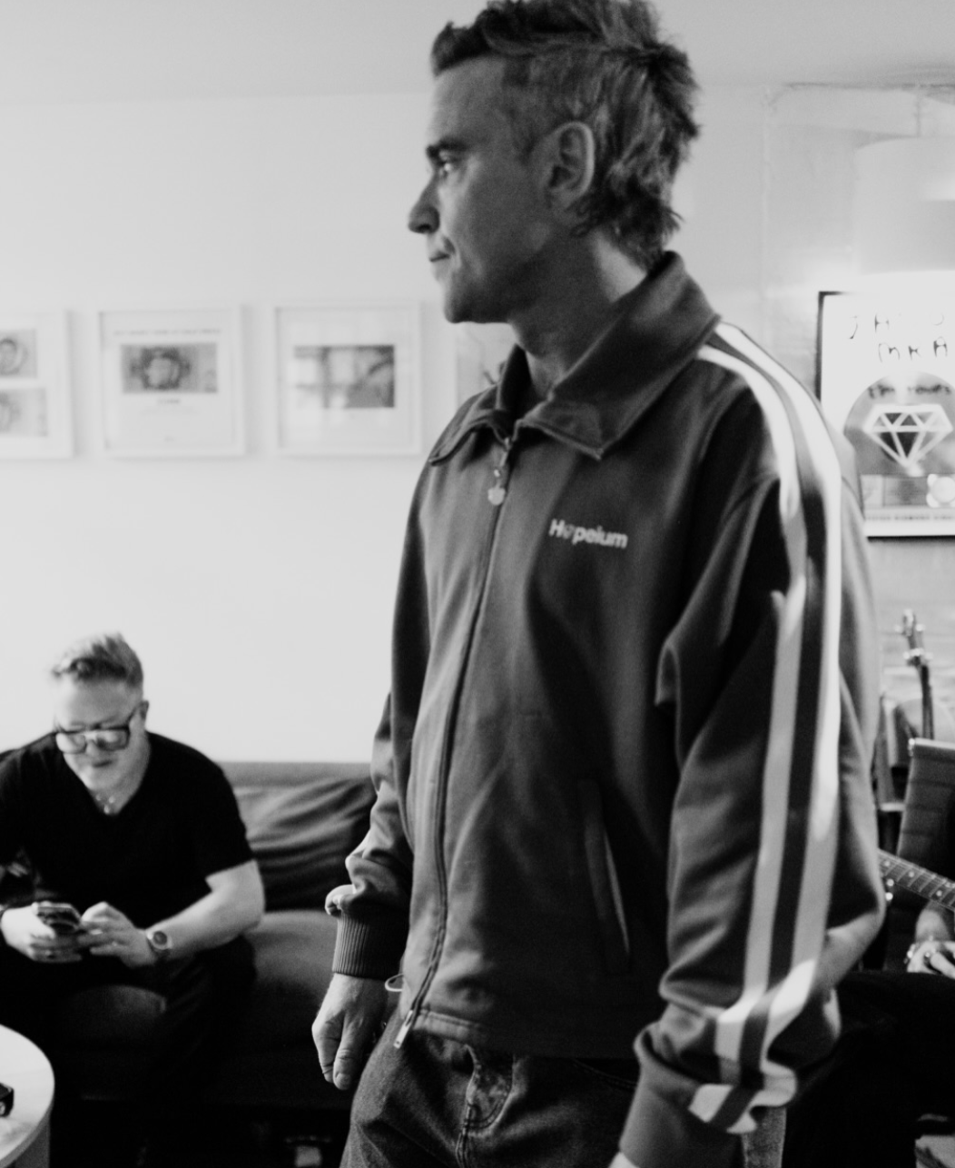 Black-and-white shot of Robbie Williams in a track jacket in a room, a man on a sofa behind him