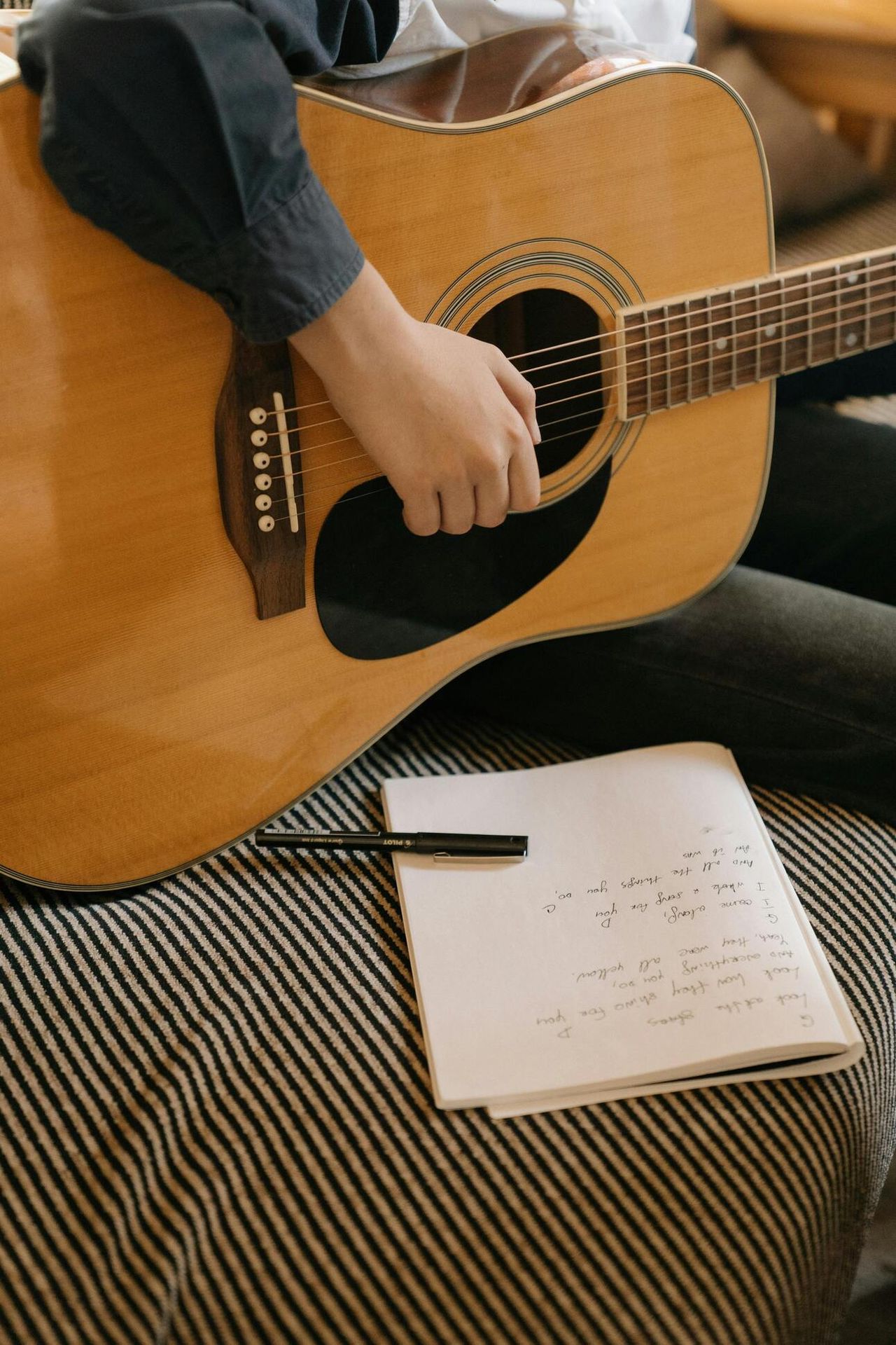 Person playing an acoustic guitar with a handwritten lyric notebook and pen beside them