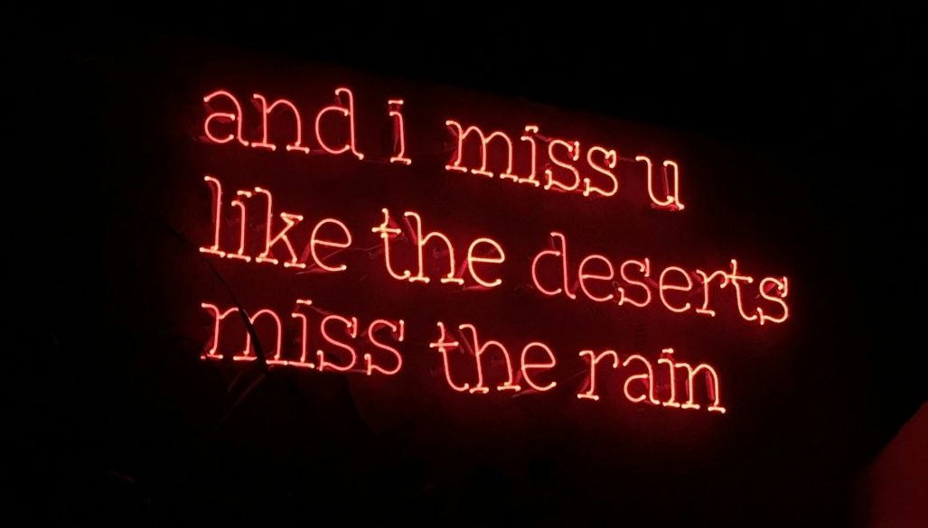 Red neon sign reading 'and i miss u like the deserts miss the rain'