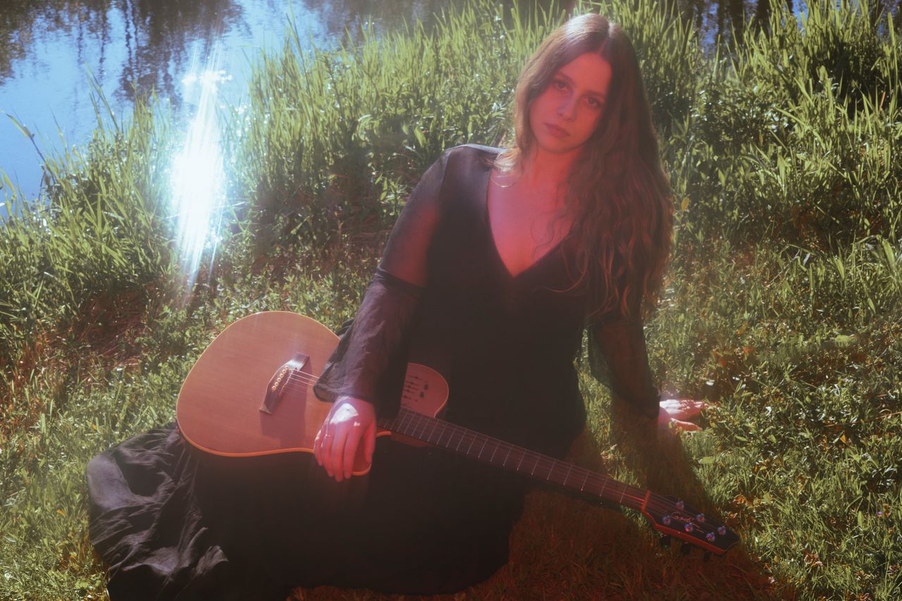 Jane Baker sitting in long grass with a guitar