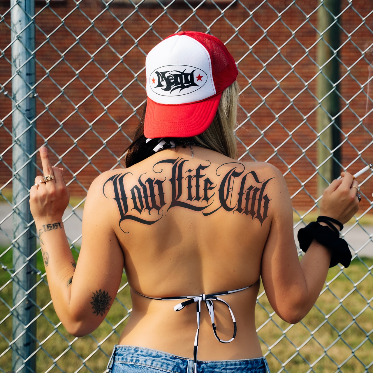 MEGG from behind in a trucker cap, a 'Low Life Club' tattoo across her back