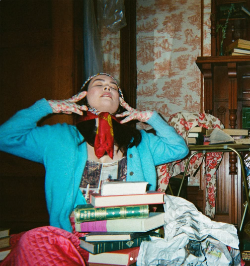 Mitski posing with eyes closed beside a stack of books in a cluttered vintage room