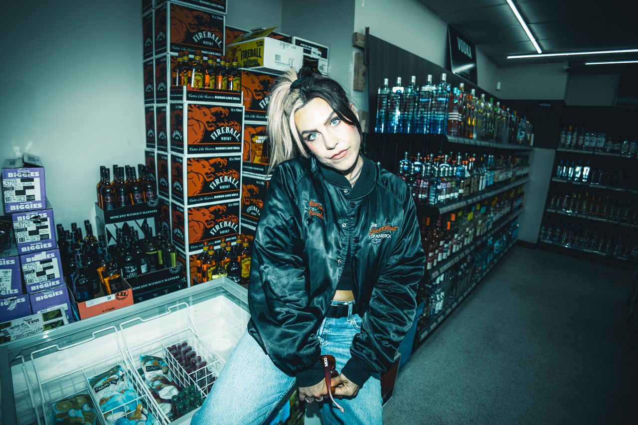 MEGG in a satin bomber jacket in a liquor store aisle stacked with Fireball boxes