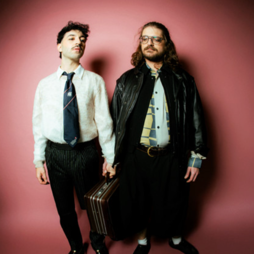 The Swiss duo against a pink wall, one holding a briefcase