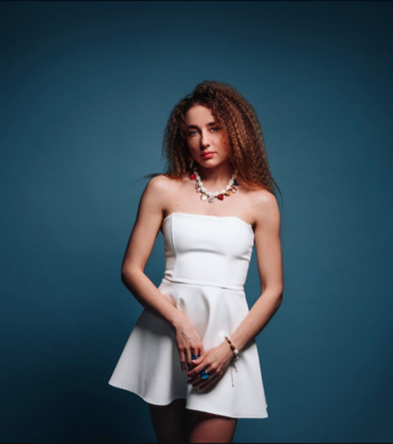 Portrait of MONARHA's singer in a white dress against a blue backdrop