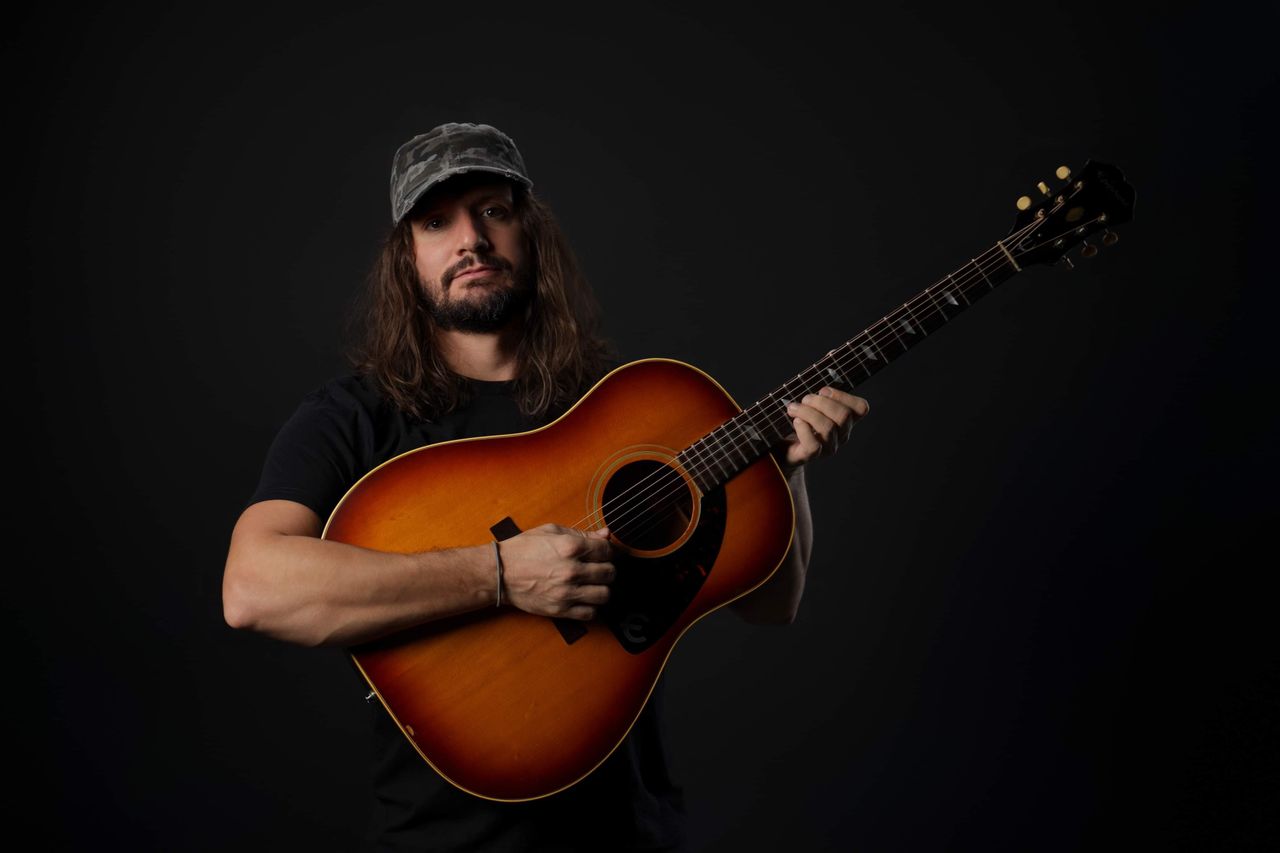 Jutt Huffman in a cap with an acoustic guitar against a black background