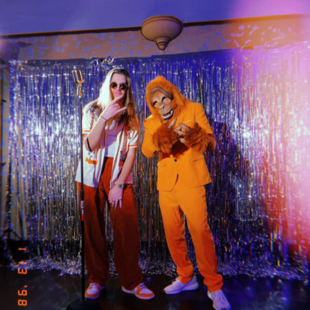 SOTB in front of a silver tinsel curtain, one in an orange orangutan mask and suit