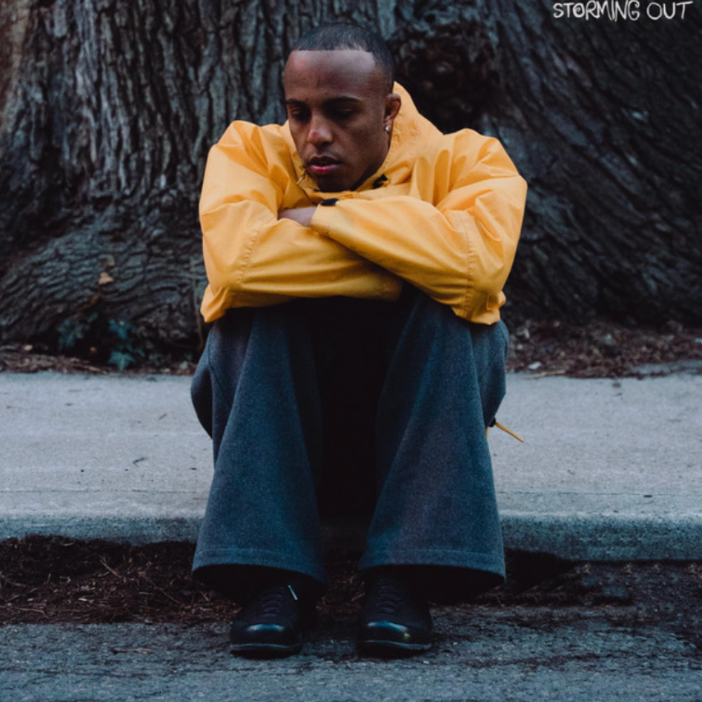 Storming Out cover - the artist in a yellow jacket sitting on a kerb