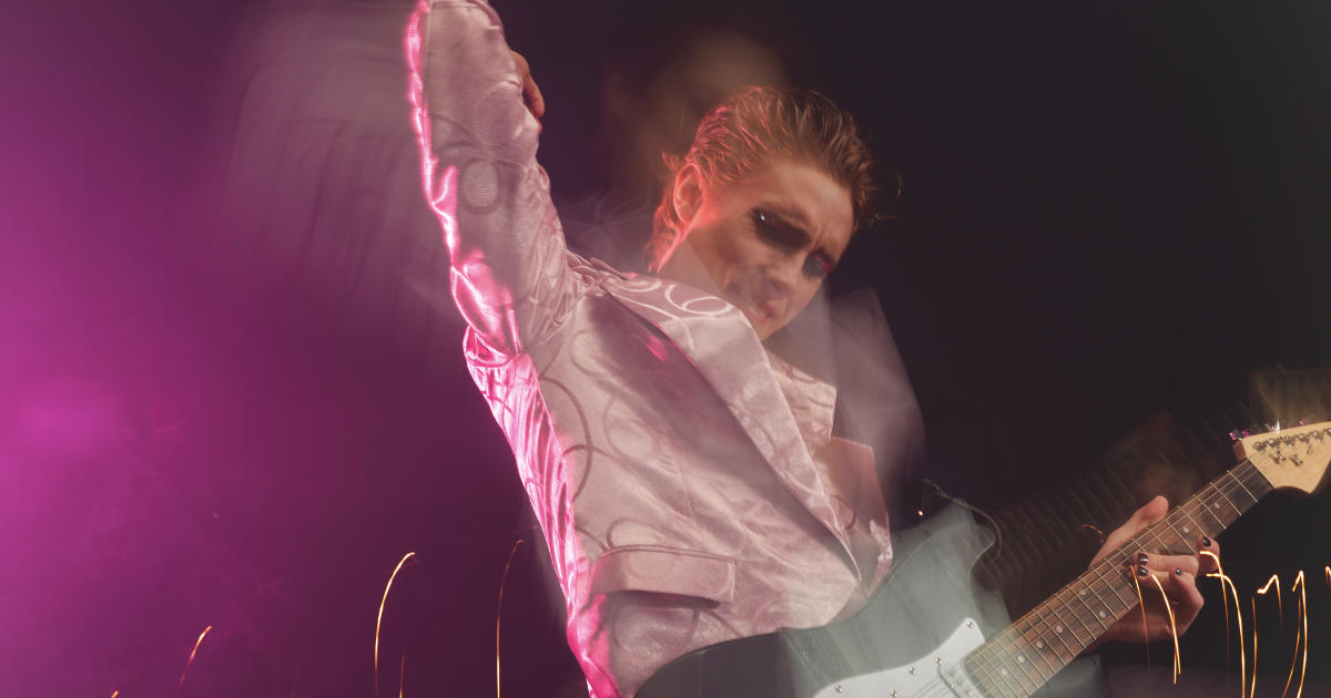 Guitarist in a pink jacket mid-performance under purple light