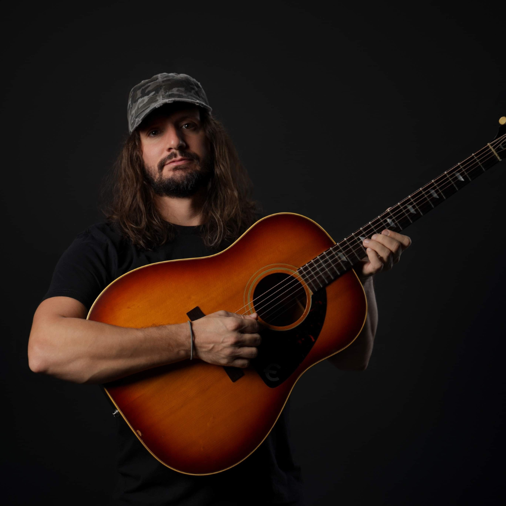 Long-haired man in a camo cap holding an acoustic guitar against black