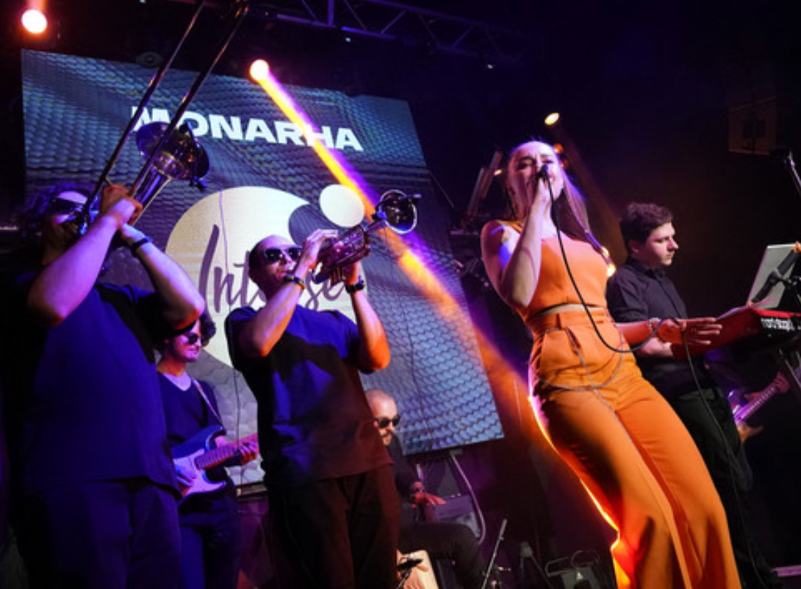 MONARHA playing live - singer in orange with a brass section and band under the MONARHA banner