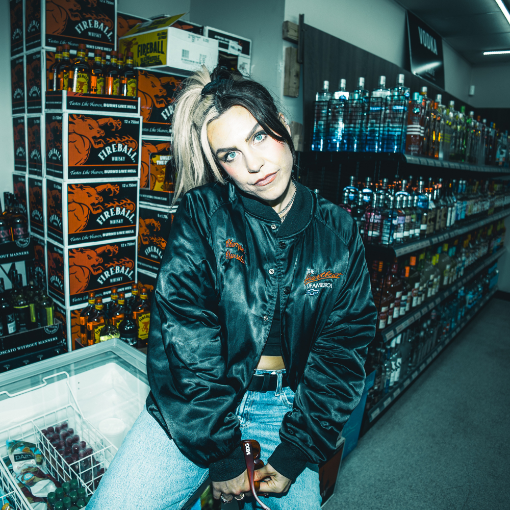 MEGG in a satin bomber jacket, posing in a liquor store aisle stacked with Fireball boxes