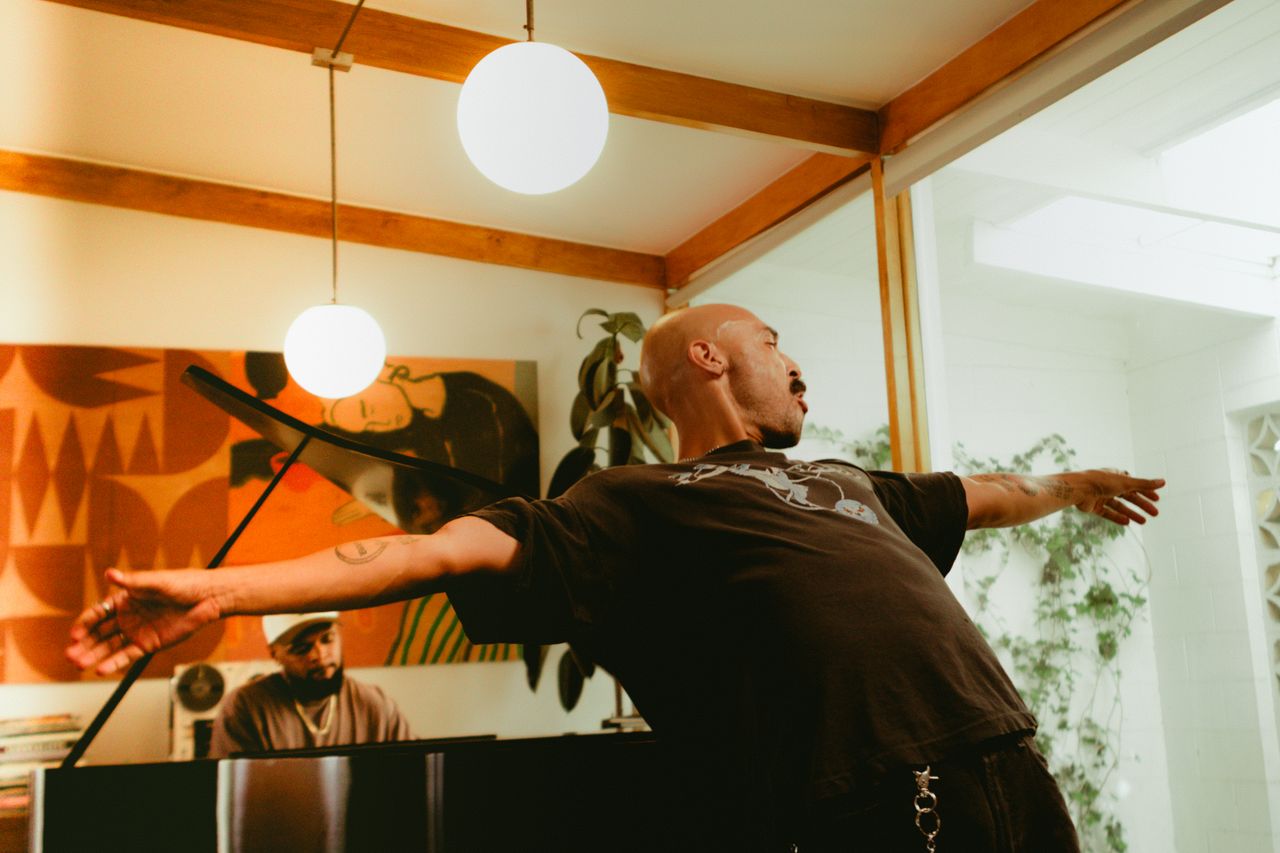 Deion Gill with arms flung wide in a sunny room, a pianist at the keys behind him